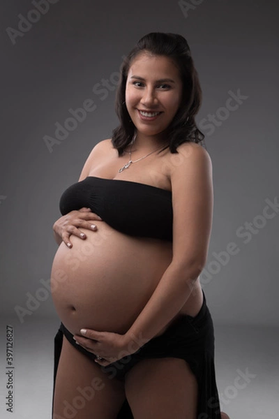 Fototapeta young latin woman smiling and touching her tummy, wearing a black top, maternity life stages, in a studio