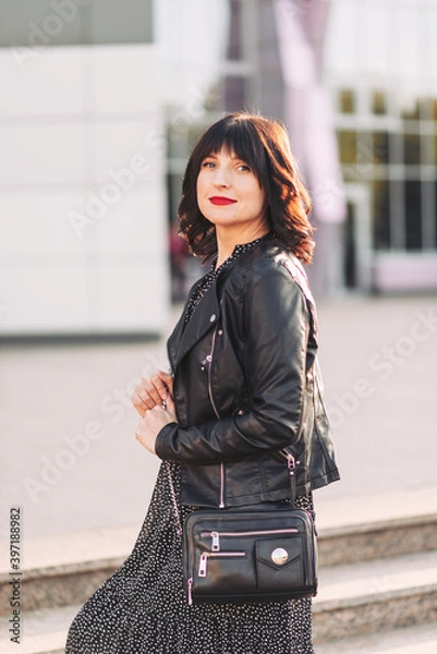 Fototapeta Portrait of a young brunette woman with red lipstick in a black leather jacket on a city street