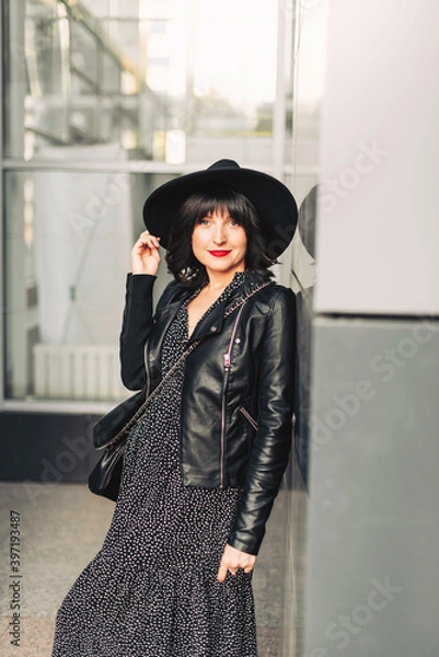 Fototapeta Young woman in a black hat, jacket and with a handbag posing on the street against a wall background