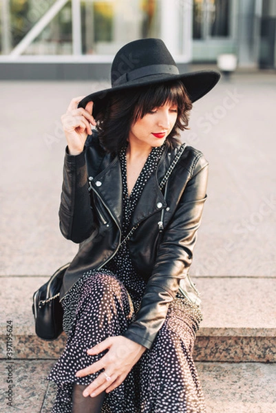 Fototapeta Hipster girl in a black hat and with red lipstick on her lips sits on the steps