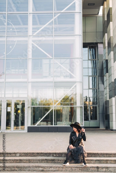 Fototapeta Hipster girl in a black hat and with red lipstick on her lips sits on the steps