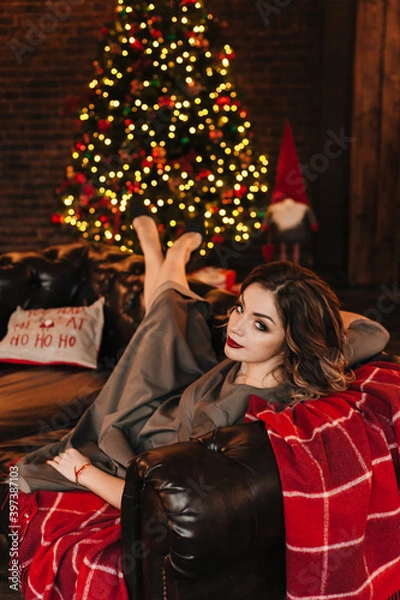 Fototapeta beautiful young girl for christmas in a happy mood against the background of a christmas tree and lights indoors