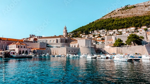 Fototapeta View of Dubrovnik from Old City Port. View of Dubrovnik ciry walls, red rooftops, and a tower with boats docked in old city harbor