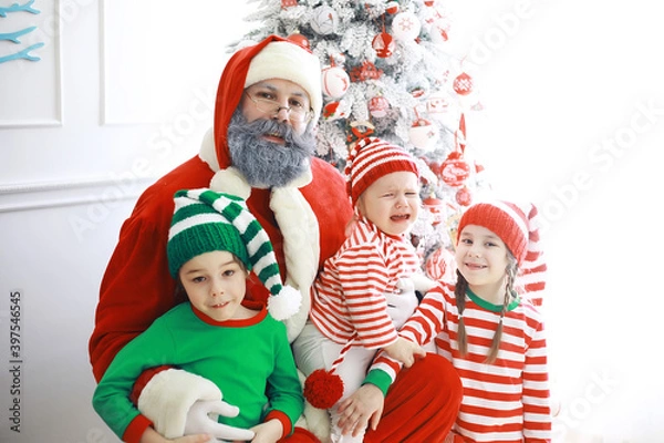 Fototapeta Santa Claus sitting with little cute elves over Christmas background. Time of miracles. Gifts from Santa Claus.