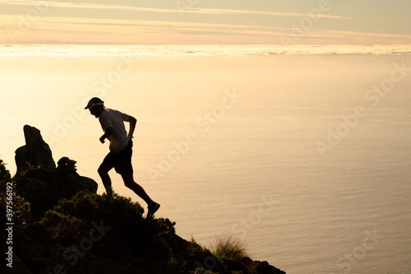 Fototapeta A runner silhouetted against the golden light of sunset over the ocean while running up a steep mountain trail.
