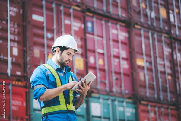 Fototapeta Man wears hardhat and reflection shirt and checking tablet with blurry metal containers in background. Concept of inventory and logistic management.