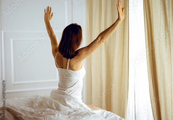 Fototapeta Woman stretching in bed after waking up, back view, entering a day happy and relaxed after good night sleep. Good morning and new day for brunette. Concept of Weekend, holidays