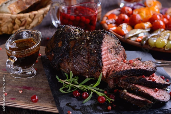 Fototapeta Appetizing roast beef with sauce and spices on a wooden table next to grilled vegetables and cranberry sauce