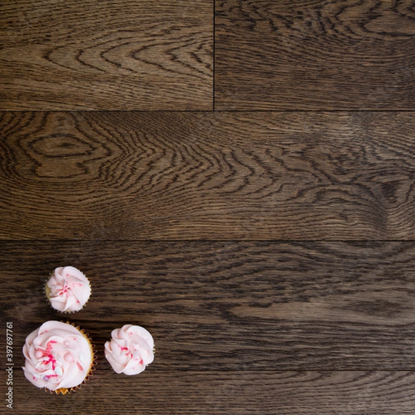 Fototapeta Three cupcakes in a corner of a wooden table; small and large cupcakes topped with pink soft icing, sprinkles and pearls