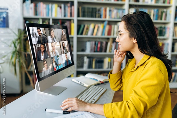 Fototapeta Online education. A female student learning online at home, focused watching at a computer monitor on which a group of other students and a teacher telling information