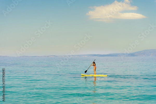 Fototapeta A single lonely girl on yellow paddle board or sup surf wearing red bikini paddling in the turquoise crystal clear water of mediterranean sea in Greece with single cloud  