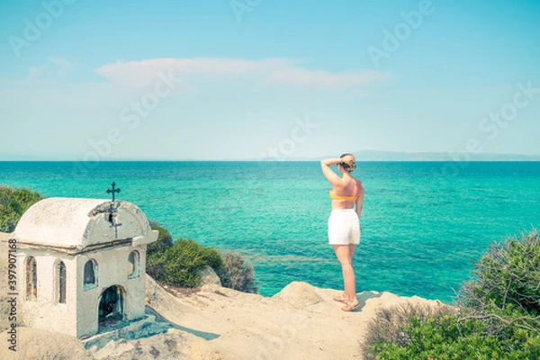 Fototapeta A girl wearing white shorts topless standing on a rock looking into the sea distance and dreaming on a sunny day near the small white temple in Greece 