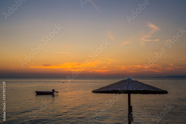 Fototapeta Sunset seascape with straw beach umbrella and a silhouette of a small boat anchored in a distance in Neos Marmaras, Greece 