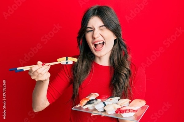 Fototapeta Young beautiful caucasian girl eating sushi using chopsticks winking looking at the camera with sexy expression, cheerful and happy face.