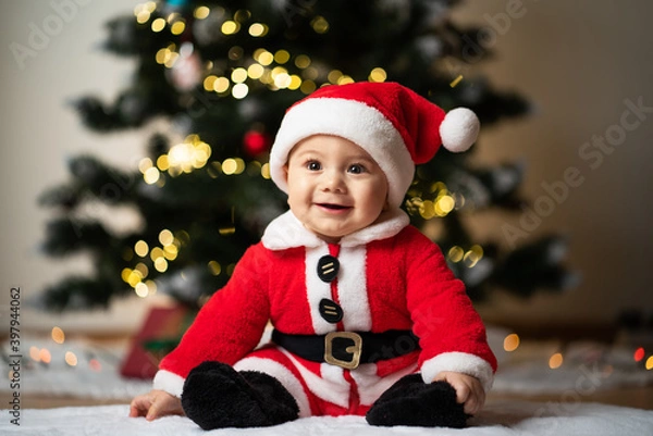 Fototapeta adorable little baby boy in a santa costume sitting on a soft fake fur in front of a christmas tree