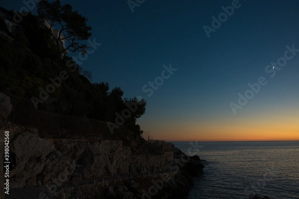 Fototapeta crépuscule sur la mer avec un dégradé de couleurs du bleu foncé à l'orange dans le ciel, un croissant de lune et le littoral dans la pénombre à gauche de l'image 
