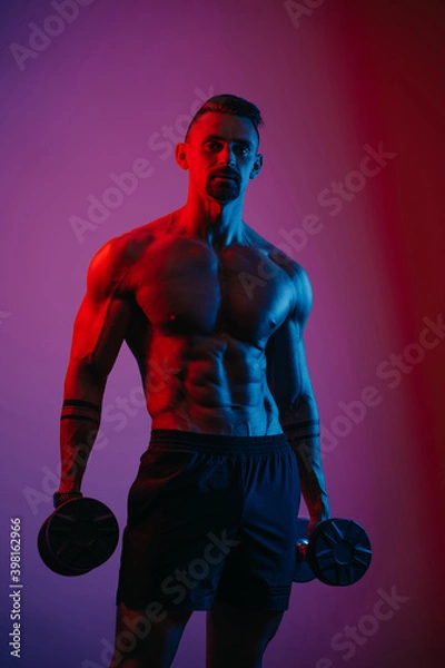 Obraz A muscular man with a beard who is posing with dumbbells under blue and red lights. An athletic guy is demonstrating his sporty torso. An athlete in shorts after biceps workout.