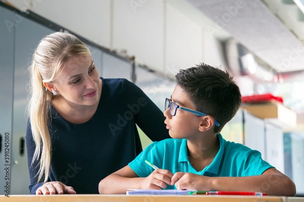 Fototapeta Cheerful school teacher giving help and support to schoolboy in class. Front view. Education or back to school concept
