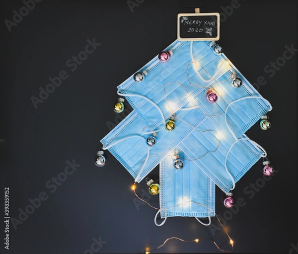 Fototapeta View of a Christmas tree made of blue surgical masks decorated with colorful Christmas baubles and Christmas lights against black chalkboar background. Christmas during pandemic or medical background.
