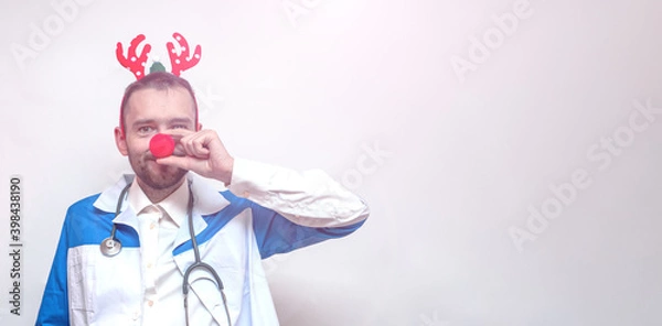 Fototapeta Young bearded doctor with a clown nose and toy deer antlers. Portrait. Place for text. Copy space
