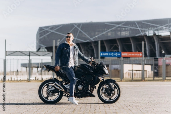 Fototapeta A man in denim clothes sitting on a black sports motorcycle against the backdrop of a large modern building