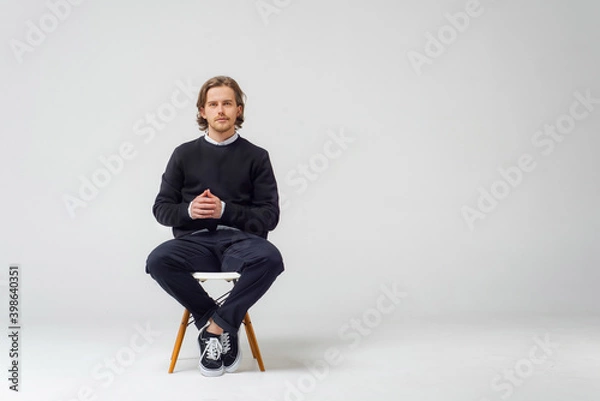 Obraz A business guy with a beard, on a white background in the studio, sits on a chair in jeans, a jacket