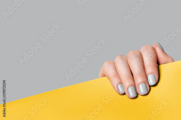 Fototapeta Female hand holds a sheet of paper and demonstrates a gray manicure. Gray, yellow background with place for text.
