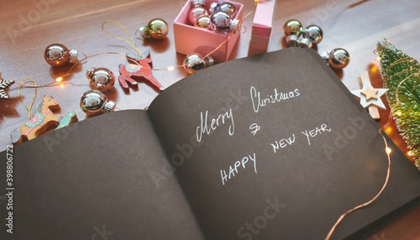 Fototapeta A black paper notebook with Merry Christmas and Happy New Year in silver letters surrounded with colorful Christmas decorations, ornaments and lights on wooden background. Happy holidays background. 