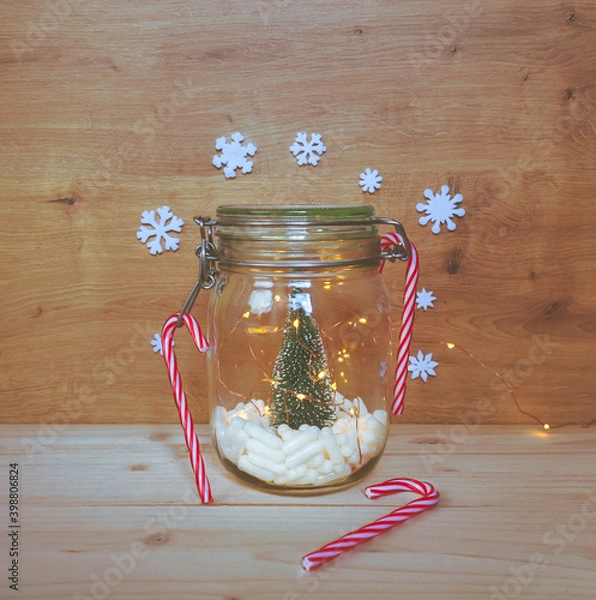 Fototapeta View of a jar filled with white capsules and a Christmas tree with lights and Christmas ornaments on wooden background with copy space. Medical or pharmacology background.