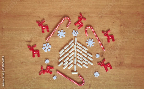 Fototapeta Christmas tree made of white capsules surrounded with red and white Christmas decorations and ornaments on wooden background with copy space. Medical, medication or pharmacy background.