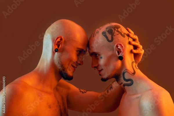 Fototapeta Portrait of young twin brothers with tattoos and piercings standing head to head, smiling at each other isolated over orange background