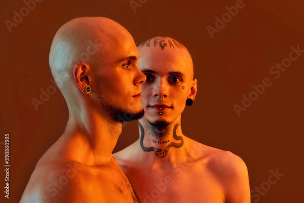 Fototapeta Front and side view of young half naked twin brothers with tattoos and piercings posing together, standing isolated over orange background