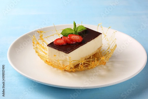 Fototapeta Dessert. Bird milk cake with strawberries and mint. Sponge cake, souffle and chocolate ganache, caramel on a white plate on a blue table. Background image, copy space, horizontal