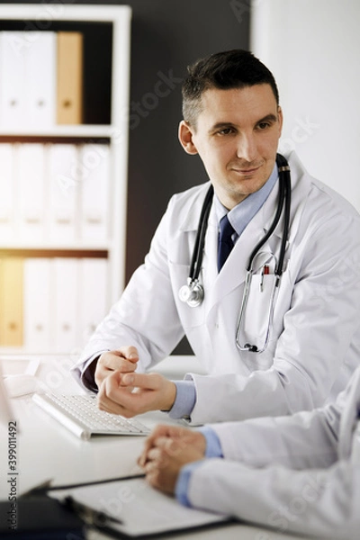 Fototapeta Two doctors colleagues discussing current disease therapy while sitting at working place in sunny clinic. Team work in hospital