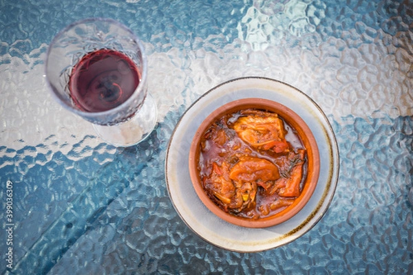 Fototapeta Georgian chicken dish with vegetables and spices in a beautiful craft plate and a glass of red wine on a glass table