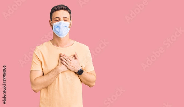 Fototapeta Handsome young man with bear wearing medical mask for coronavirus smiling with hands on chest with closed eyes and grateful gesture on face. health concept.