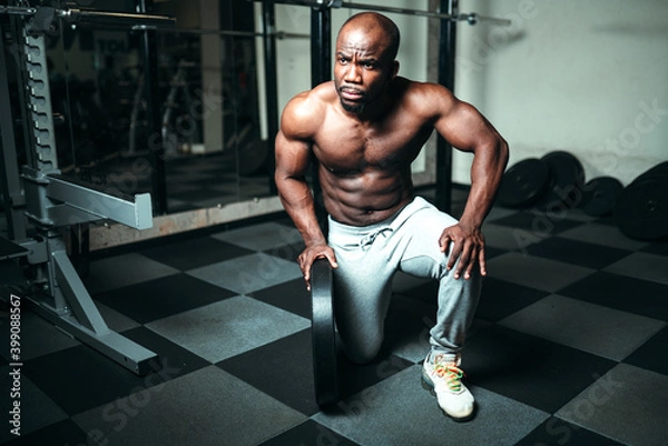 Fototapeta Male african american powerlifting trainer sitting on one knee on the floor in the gym with a barbell disc in hand