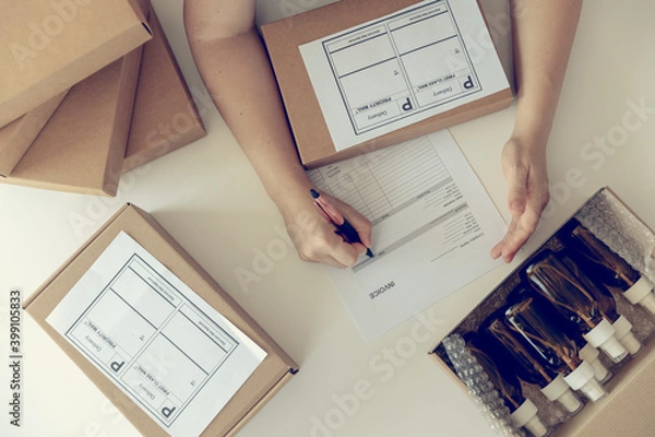 Fototapeta Close-up of woman`s hands on the white desk with carton boxes and signing the parcel with dark glass bottles for delivery. Online working concept.