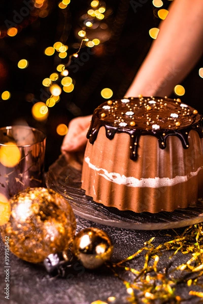 Fototapeta Holding tasty Three Chocolates mousse cake with ganache in Christmas and New Year decorations. Chocolate cake with layers. Bokeh lights background. Vertical orientation. Christmas Eve. Low Key.