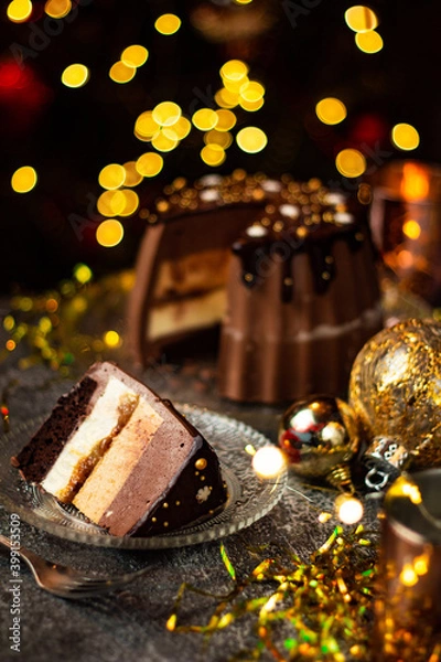 Fototapeta A piece of tasty Three Chocolates mousse cake with ganache in Christmas and New Year decorations. Chocolate cake with layers. Bokeh lights background. Vertical orientation. Christmas Eve. Low Key.