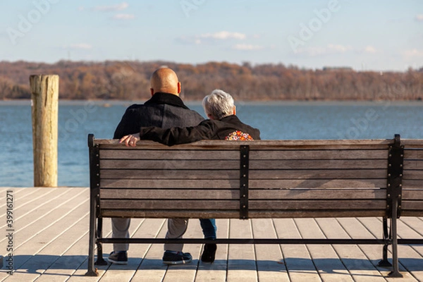 Fototapeta an elderly caucasian woman with short gray hair and a younger African American man with shaved head are sitting on a bench on a pier looking at sea. Image for interracial couple with age difference.