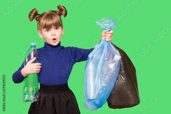 Fototapeta Little ecologist girl with confused funny face holding trash bags with plastic bottles isolated on green background