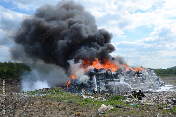 Fototapeta A group of firefighters extinguishes huge landfill fire with flames and black smoke in the background