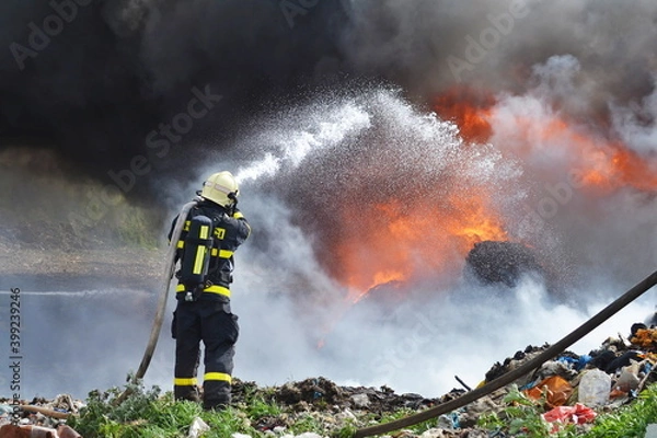 Fototapeta A fireman extinguishes huge landfill fire with flames and black smoke in the background