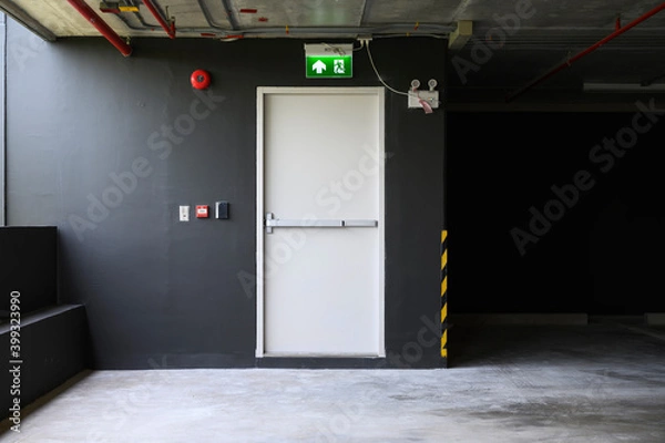 Fototapeta A fire exit with the sign and red alarm bell above the white door on the parking building.