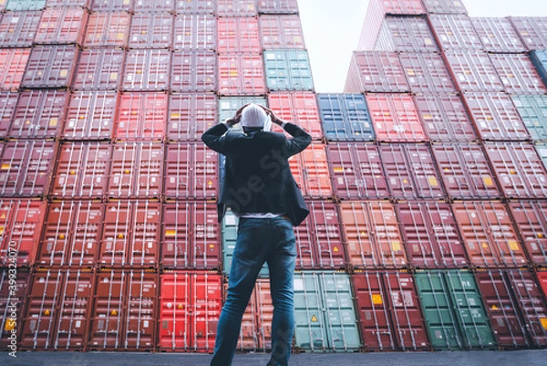 Fototapeta Worker or foreman standing in front of stacked cargo containers and hands hold his head wears white hardhat and hold face mask ack like a shock or surprise. Difficulties in transport during Covid-19.