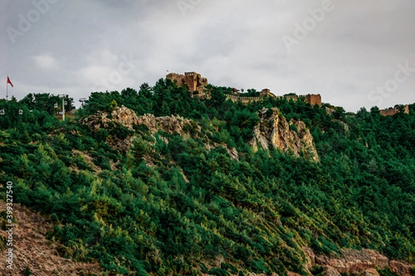 Fototapeta Alanya Castle (Turkey) on the top of a mountain among a green forest - bottom view. Ancient fortress with a tower and stone walls among green trees on a hill in a summer-autumn cloudy day