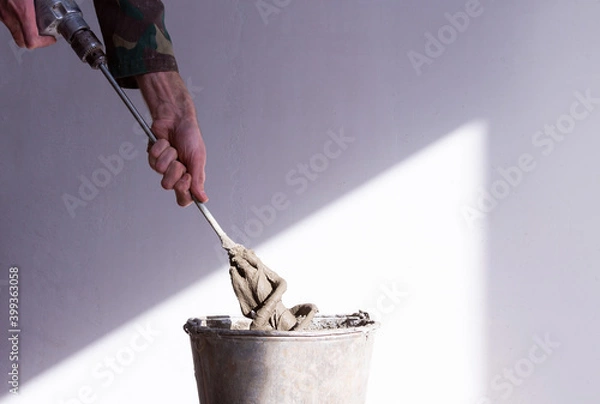 Obraz stirrer in the hand of a worker in a cement mortar and an old rusty bucket close up