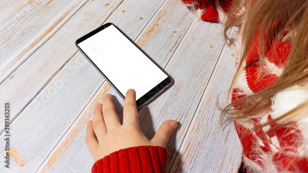 Fototapeta The child is sitting at a table with a phone with a white screen. A girl with a phone layout. In new year's clothing. New Year