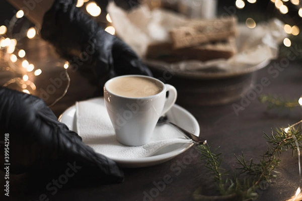 Fototapeta A waiter in medical gloves puts a cup of espresso on the table in coffee shop, Christmas bokeh lights. Selective focus